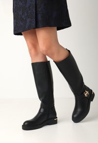 Botas de cuero negro hasta la rodilla con puntera redondeada, cierre de cremallera y un acento de logo dorado en la correa del tobillo. Acabado mate texturizado.