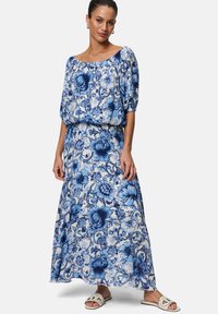 Robe maxi en tissu léger avec des motifs floraux bleus. Encolure dénudée, manches bouffantes et silhouette fluide. Sandales avec une sangle texturée.
