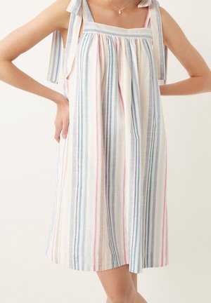 Femme portant une robe sans manches longueur genou avec des rayures verticales pastel et des bretelles nouées aux épaules, debout les mains sur les hanches.