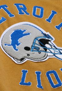 Detroit Lions Grafik auf einem senffarbenen Hintergrund. Zeigt einen grauen Helm mit einer blauen Löwensilhouette und blauem Text, der den Teamnamen umreißt.