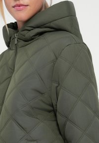 Giubbotto imbottito trapuntato verde oliva con cappuccio, chiusura frontale con zip e motivo a rombo. Leggero con una texture morbida.