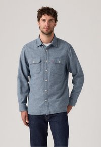 Blue patterned button-up shirt made of a soft fabric, featuring two chest pockets and a pointed collar. Worn with dark jeans.