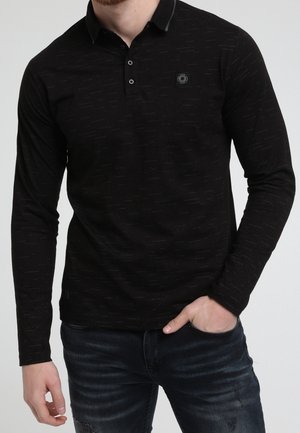 Mann trägt schwarzes langärmeliges Poloshirt mit feinem Muster und dunkle Jeans, mit linker Hand in der Tasche und rechter Hand locker an der Seite.