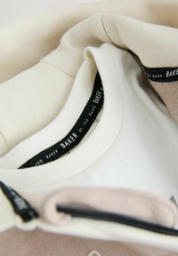 Beżowa i kremowa warstwowa tkanina o miękkiej fakturze, z czarną metką "TED BAKER" przy dekolcie. Widoczny detal zamka błyskawicznego.