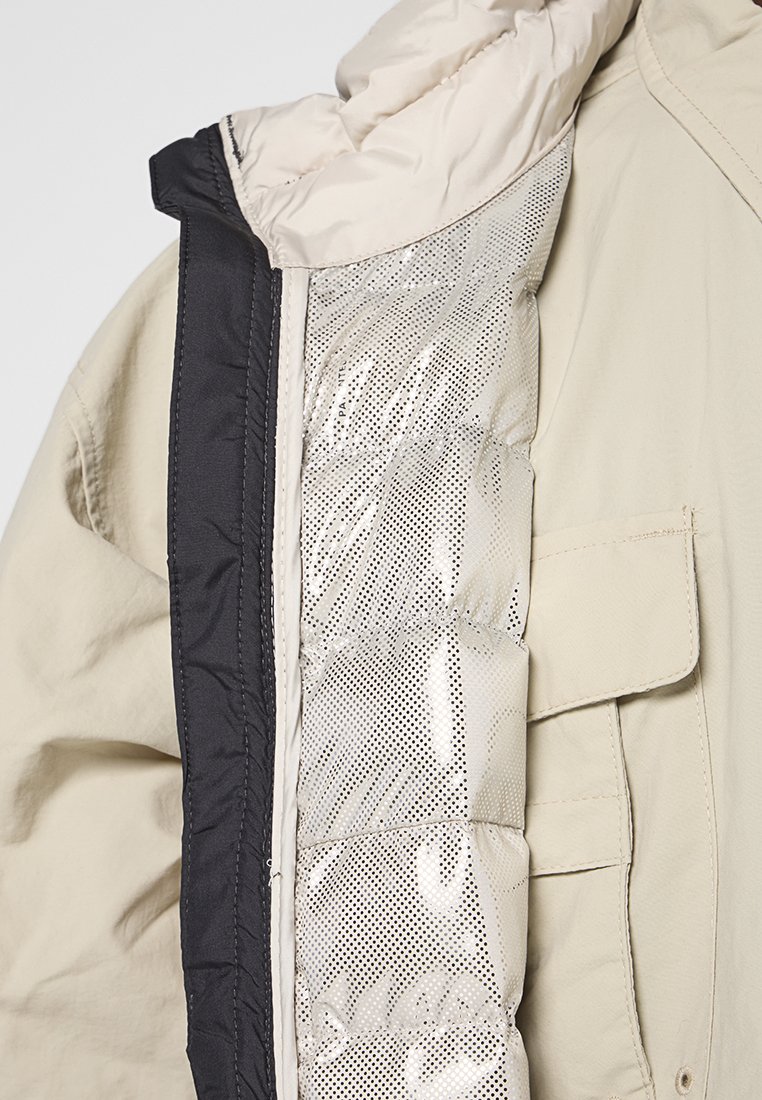 Giacca beige con rifinitura nera sulla zip e foderatura interna argentata, caratterizzata da piccole perforazioni e un delicato motivo testuale.