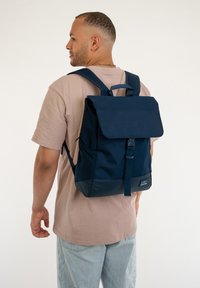 Mochila azul marino con cierre de solapa, correas ajustables, tela texturizada y un hebilla en el frente, con bolsillos laterales y un parche de logo.