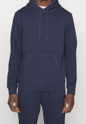 Homme portant un sweat à capuche bleu foncé avec poche avant et cordon de serrage à la capuche, associé à un pantalon de survêtement bleu assorti, sur un fond uni.
