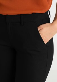 Černé ušité kalhoty s hladkou texturou, vybavené bočními kapsami a slim fit střihem. Nošeny s oranžovou blůzou, která ukazuje detaily poutka na pásek.