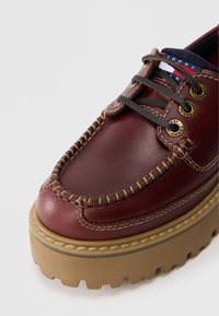 Mocasin din piele burgundy cu talpă groasă din cauciuc bej, șireturi maro prin ochiuri din alamă și un mic etichetă cu logo-ul Tommy Jeans pe limbă.