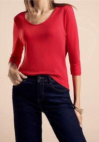 Rotes Langarmoberteil mit Rundhalsausschnitt, kombiniert mit dunkelblauen Jeans, die über Vorderaschen und sichtbare Nähte verfügen.