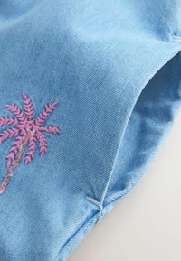 Tissu en denim bleu clair avec un petit détail brodé d'un palmier rose et des surpiqûres visibles sur le bord.