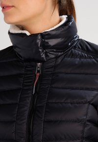 Veste matelassée noire avec un col haut, dotée d'une fermeture éclair et d'une doublure en tissu texturé contrastant. Les coutures forment un motif matelassé.