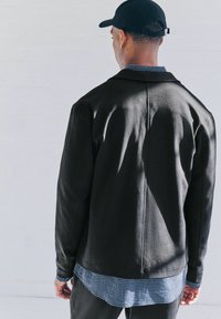 Zwarte jas met een gladde textuur, gevouwen kraag en achternaaddetail, gedragen over een blauw-wit geruit shirt.