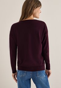 Vrouw die naar de zijkant glimlacht, gekleed in een donkerpaars langemouwen trui en blauwe jeans, staand tegen een effen lichte achtergrond.