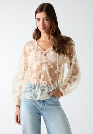 Mujer joven con cabello largo y castaño que lleva una blusa blanca transparente bordada con flores y vaqueros azul claro, posando con una mano en el bolsillo.