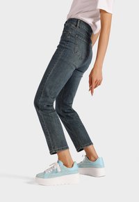 Jeans blu scuro con vestibilità slim e lunghezza cropped, abbinati a sneakers platform azzurri chiari con una superficie texturizzata e suole bianche.