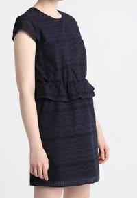 Nainen pukeutuneena tummansiniseen lyhythihaiseen mekkoon, jossa on silmukkalace-kuvioita ja kerätty peplum-vyötärö, seisoo yksinkertaista taustaa vasten.