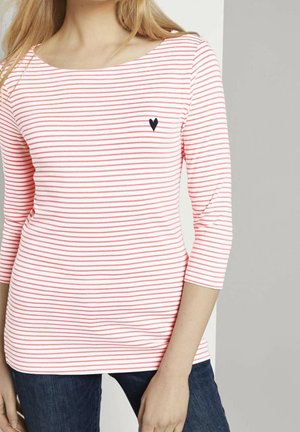 Kvinde iført en hvid langærmet skjorte med tynde røde vandrette striber og et lille sort hjerteemblem på brystet, kombineret med mørke jeans.