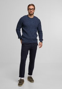 Granatowy sweter z dzianiny o fakturowanym wzorze, okrągły dekolt; zestawiony z ciemnymi spodniami i brązowymi zamszowymi butami. Model stoi z rękami w kieszeniach.