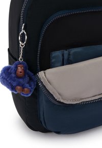 Sac à dos noir et bleu marine avec une poche grise clair. Il est doté d'un porte-clés en forme de singe en peluche bleu avec des accents marron attaché à la fermeture éclair.