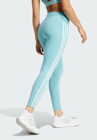 Legíny v aqua modrej farbe s bielymi bočnými pruhmi, vysoký pás a hladká elastická textúra. Kombinované s bielymi športovými topánkami.