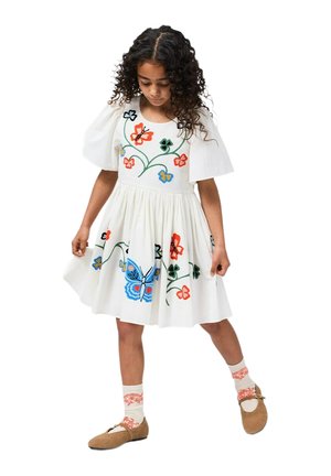 Robe blanche avec manches courtes bouffantes, ornée de broderies colorées de fleurs et de papillons. Associée à des chaussures beige et des chaussettes à motifs.