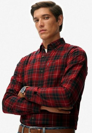 Red and black plaid flannel shirt with long sleeves, buttoned collar, and front pocket detail, worn with a brown leather belt.