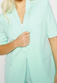 Mintgroene blazer met korte mouwen, enkele knoopsluiting en voorzakken. Gladde textuur met een gestructureerde silhouet.