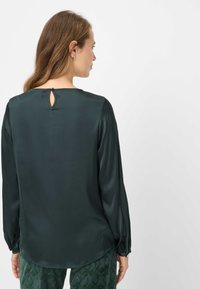 Mørkegrøn, silkeagtig bluse med lange ærmer, rundet halsudskæring og en lille nøglehulsåbning bagpå. Har sammensyede ærmekanter.