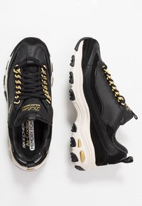 Svarta lädersneakers med guldaccenter, med en tjock sula med beige sektioner och snörningsdesign. Inkluderar "Skechers"-branding.