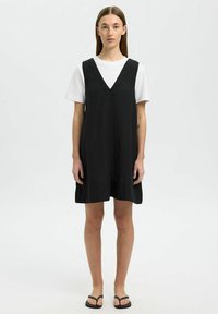 Zwarte mouwloze jurk met een V-halsontwerp, gemaakt van lichtgewicht stof, met een losse pasvorm en subtiele textuur. Draag het over een wit T-shirt.