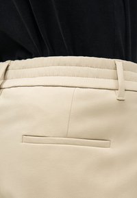 Nahaufnahme von beigen, maßgeschneiderten Hosen mit Gürtelschlaufen und einer geschlitzten Gesäßtasche, getragen mit einem vorne in die Hose gesteckten schwarzen Oberteil.
