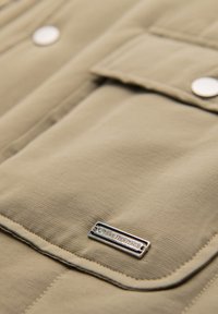 Primer plano de un bolsillo de tela beige con solapa, botones de presión metálicos y una etiqueta metálica rectangular que dice "Oxbow Heritage".