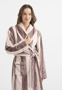 Kobieta w miękkim, prążkowanym szlafroku w odcieniach brązu i kremu, trzymająca kołnierz jedną ręką na białym tle.