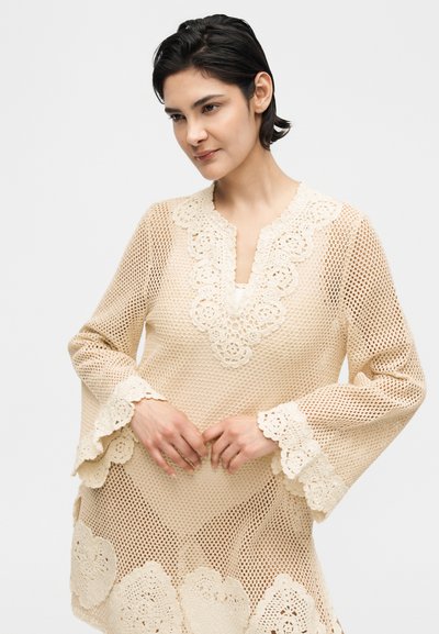 Νεαρή γυναίκα με μπεζ πλεκτό φόρεμα με λεπτομέρειες από δαντέλα με λουλούδια στα μανίκια, το ντεκολτέ και το τελείωμα, ποζάρει μπροστά από απλό φόντο.