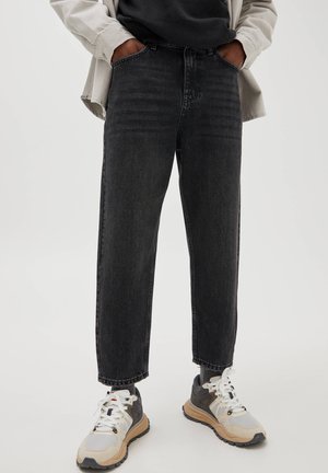 Person mit weiten schwarzen Jeans, beiger Jacke, schwarzem Shirt und beigen Sneakers steht mit den Händen in den Taschen vor schlichtem Hintergrund.