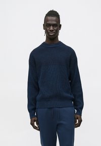 Fonott hajú férfi, sötétkék, mint�ás pulóvert és hozzá illő sötétkék nadrágot visel, sima, világos háttér előtt.