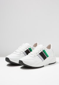 Białe sneakersy z zielonymi i czarnymi akcentami, charakteryzujące się grubą podeszwą, warstwowym designem i teksturowanymi panelami z siateczki. Szczegóły sznurowadeł i pasków.