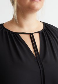 Schwarze Bluse mit plissiertem Design, ausgestattet mit einem Schlüsselloch-Ausschnitt und schwarzem Bandverschluss. Hergestellt aus weichem, glattem Stoff mit einer lockeren Passform.