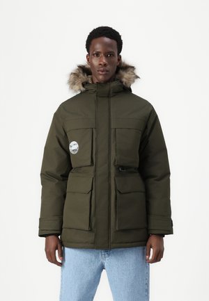 Olive green parka with fur-trimmed hood, large front pockets, and logo patch. Thick fabric with a matte finish, paired with light blue jeans.