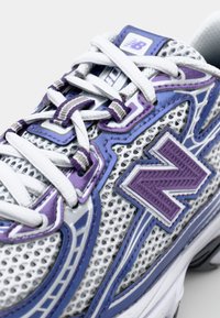 Sapatilha de malha branca com detalhes em roxo e sola de borracha. Apresenta uma ponta arredondada, atacadores e um logótipo "N" texturizado. Elementos de design geométrico.