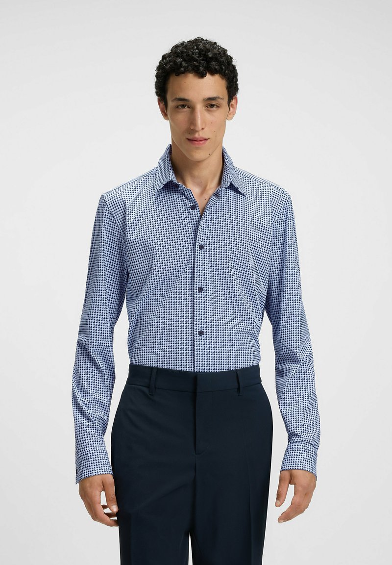 La camicia a maniche lunghe a quadri blu e bianchi presenta un colletto a punta e bottoni frontali, abbinata a pantaloni eleganti scuri. Texture del tessuto liscia.