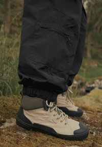 Beige High-Top-Stiefel mit schwarzen Gummisohlen, strukturiertem Obermaterial, grünen Akzenten und gemusterten Ösen. Schwarze Hosen mit elastischen Bündchen.