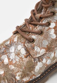 Bota de cuero marrón con superficie texturizada que presenta detalles metálicos y un patrón manchado. Tiene cordones marrones y ojales de metal.