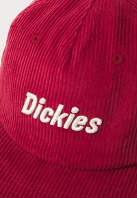 Casquette en velours côtelé rouge avec le texte "Dickies" brodé en blanc à l'avant. Présente des rayures verticales texturées et une visière incurvée.