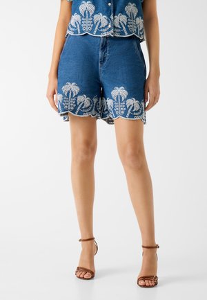 Frau trägt blaue Jeans-Shorts mit weißen bestickten Palmenmustern und braune Sandaletten mit Absatz und Knöchelriemen auf weißem Hintergrund.