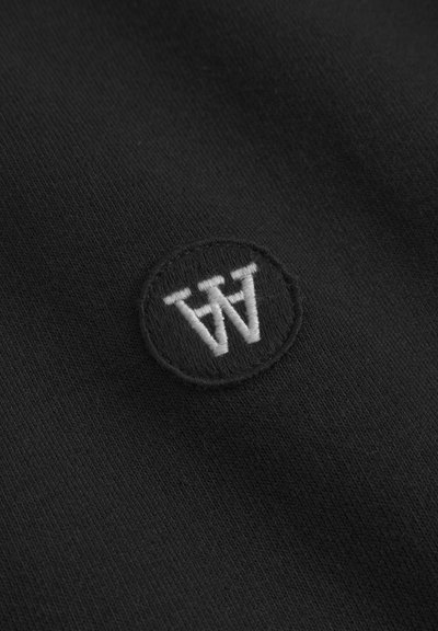 Schwarzer Stoff mit einer strukturierten Oberfläche, auf dem ein kreisförmiges, in Weiß und Grau gesticktes Logo zu sehen ist, bestehend aus stilisierten Buchstaben "W" und "M."
