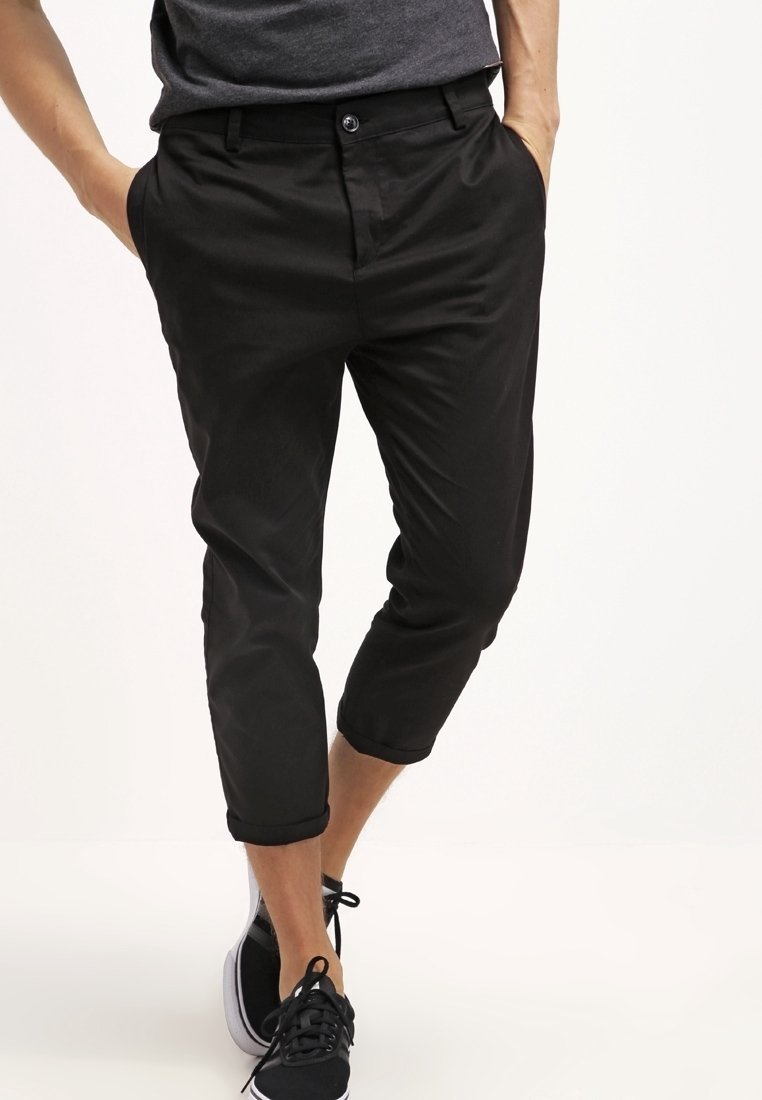 Pantalon noir coupé court avec une coupe ajustée, doté d'une fermeture à bouton, de poches latérales et d'une texture lisse. Porté avec des baskets noires.