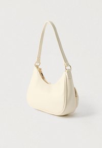 Petit sac à bandoulière beige avec une seule lanière, forme incurvée et détail de fermeture éclair dorée sur fond neutre.