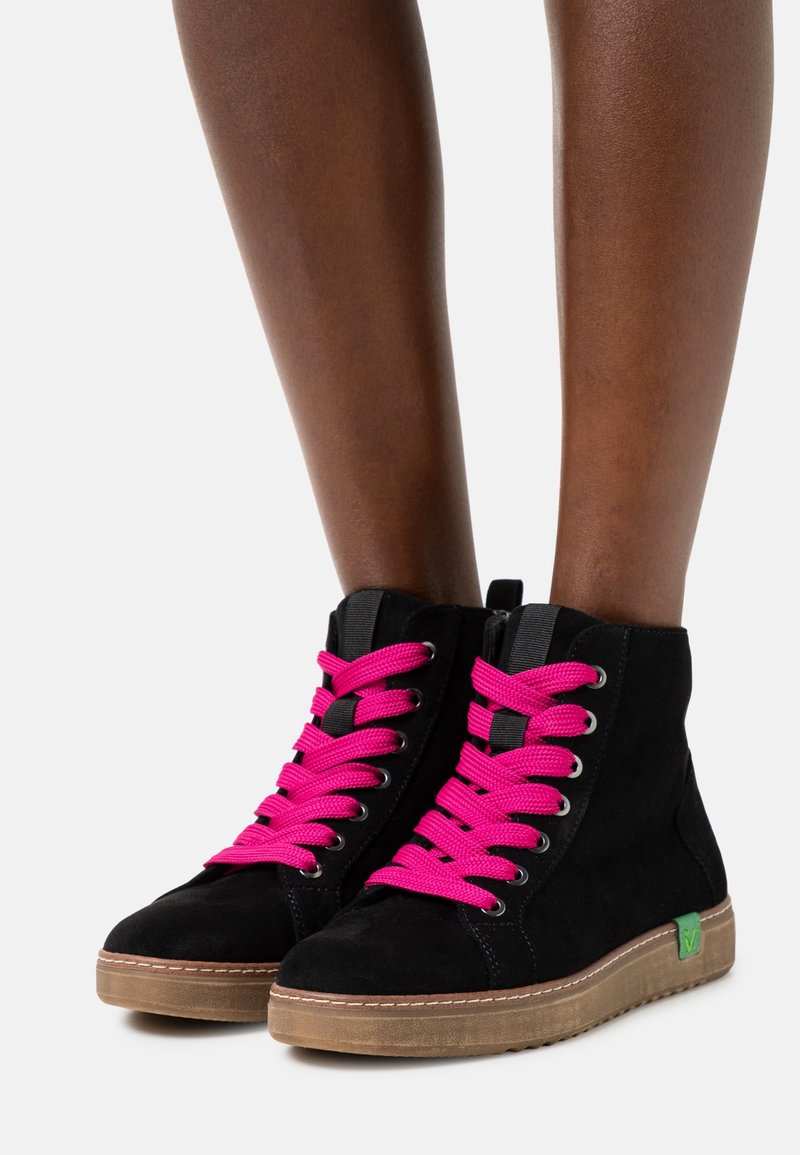 Baskets montantes noires avec des lacets rose vif et des semelles en gomme, portées sur des jambes brun foncé, vues de face et de côté.